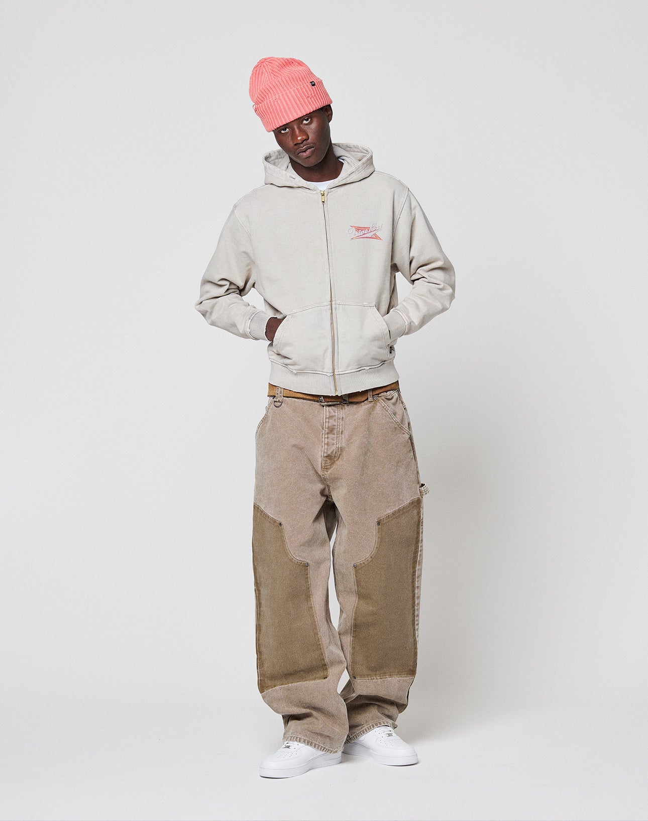 A person wears the LIVE FAST DIE YOUNG LFDY Corp. Hooded Zip in light gray, loose tan work pants with unique wash effect, a bright pink beanie, and white sneakers, standing against a plain white background.