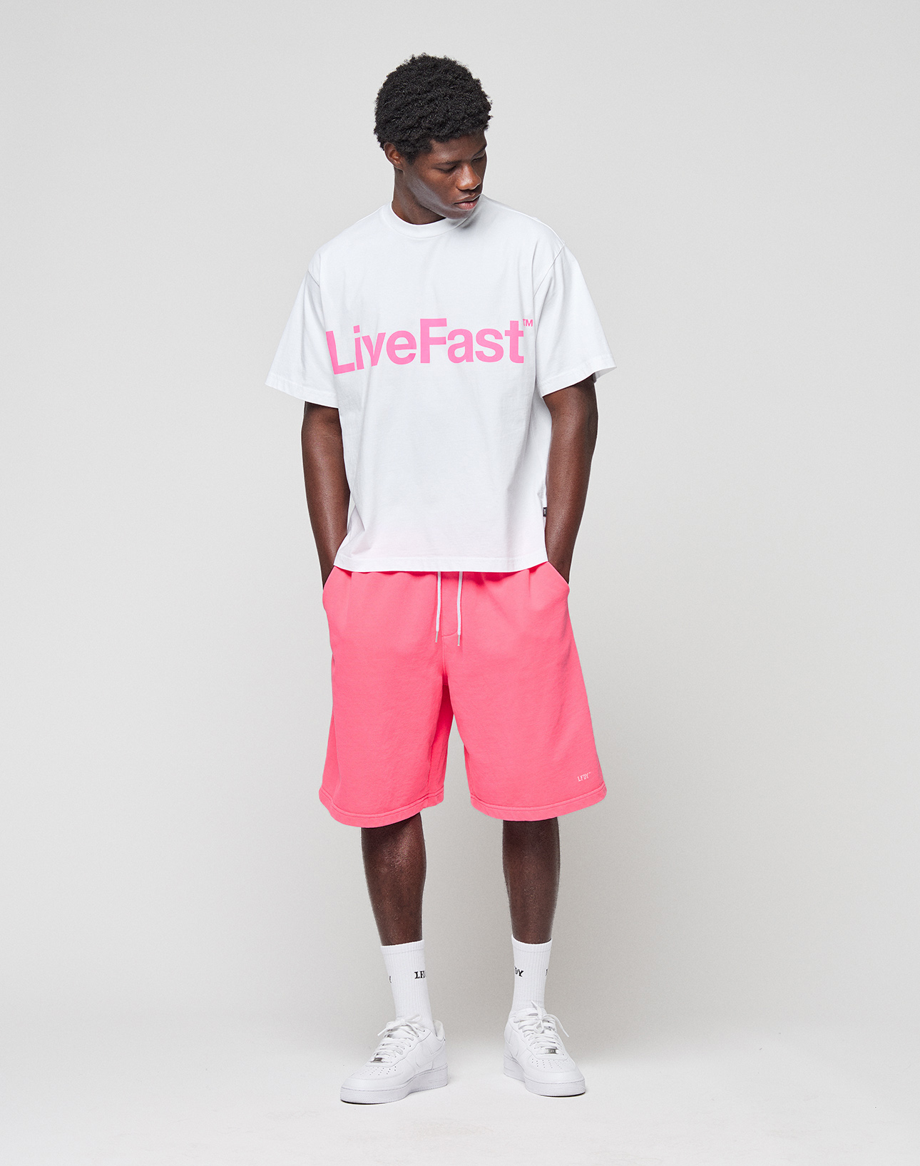 A man stands against a plain background wearing a white LIVE FAST DIE YOUNG Logo-Print t-shirt with "LiveFast" in bold pink, Basic Shorts with elastic waistband in pink, white socks, and white sneakers. He looks down with hands in his shorts pockets.