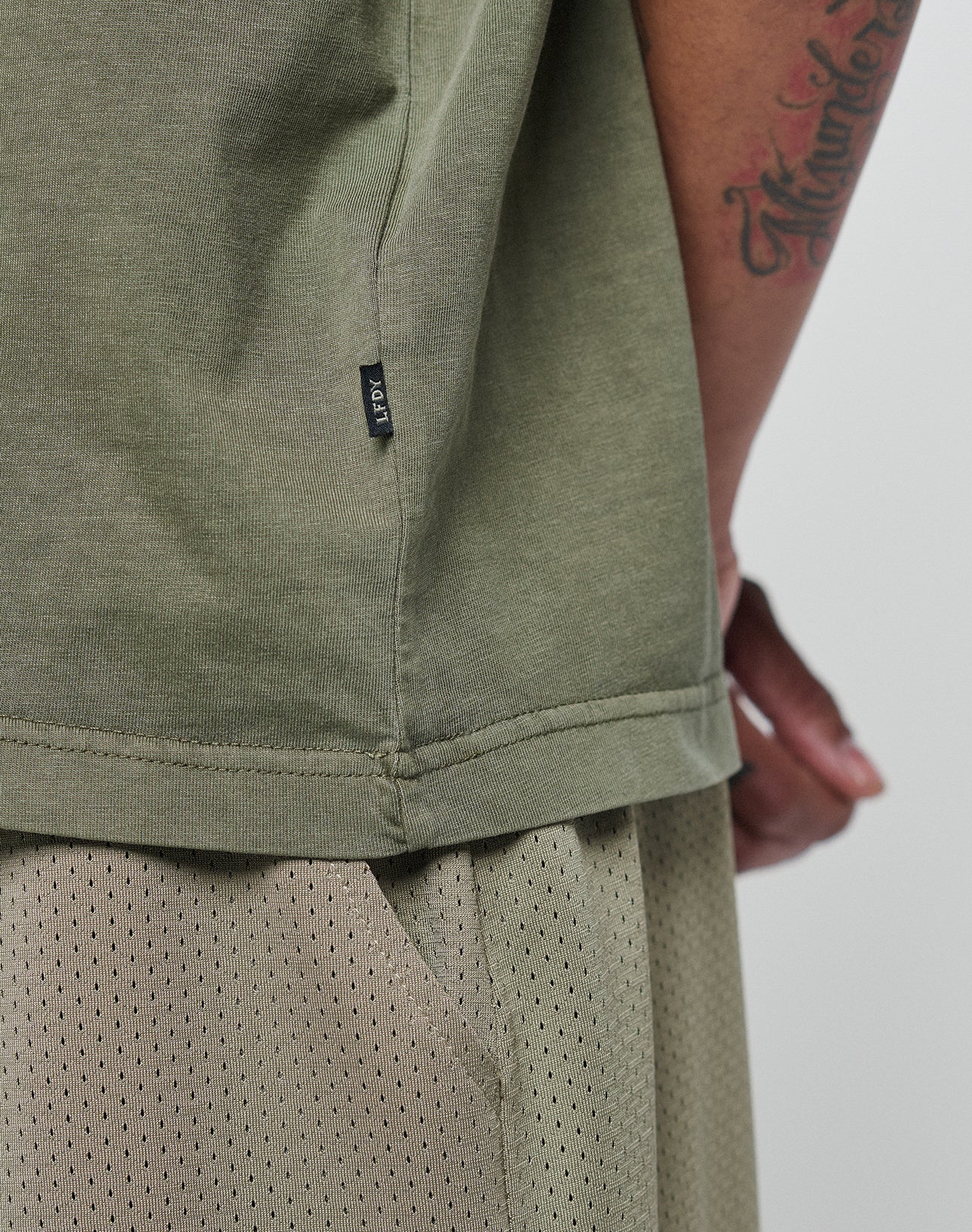 A close-up shows a person in the LIVE FAST DIE YOUNG Basic +49 Chest Tee in olive green with a visible tattoo and clothing tag, paired with beige mesh shorts.