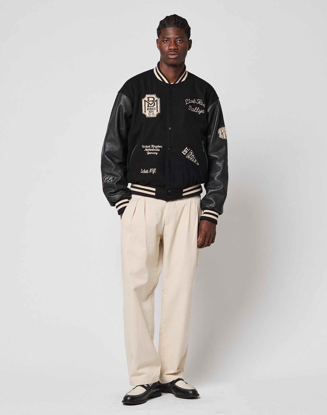 A man stands against a white background wearing the limited edition LIVE FAST DIE YOUNG x Schott NYC Varsity Jacket with white details and patches, cream pleated trousers, and black-and-white dress shoes.
