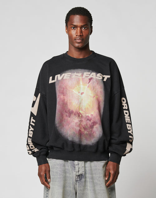 A man wears the LFDY Angel Sweater by LIVE FAST DIE YOUNG—an oversized black Baumwolle sweatshirt with LIVE FAST text above a vibrant graphic and bold OR DIE BY IT text on the sleeves, standing against a plain light background.