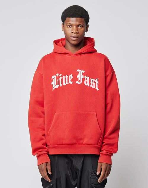 A person faces forward in the LIVE FAST DIE YOUNG Live Fast Fractured Hooded—an oversize fit red hoodie with doppellagige Kapuze and bold white Live Fast print, made from Baumwolle Polyester, paired with black pants on a light gray background.
