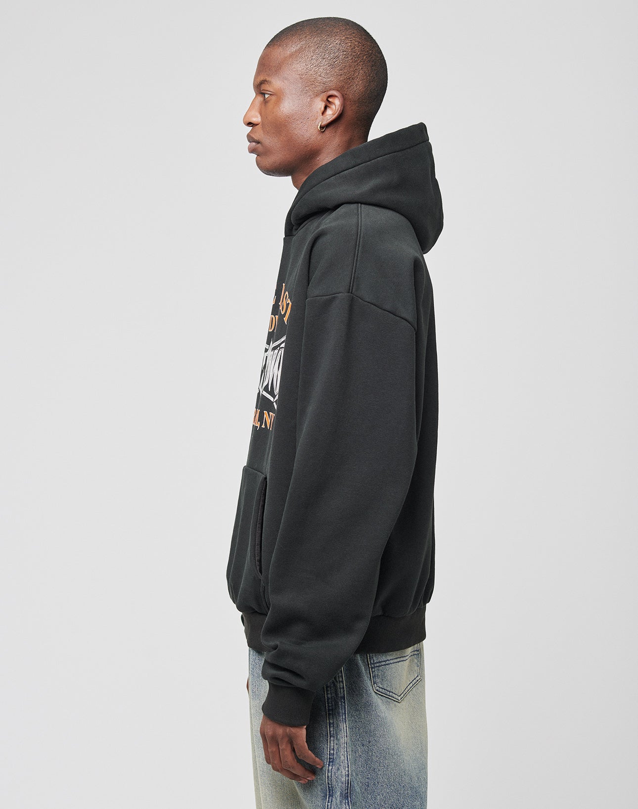 A person in profile against a plain light gray background wears the LIVE FAST DIE YOUNG Live Fast Tribal Hooded sweatshirt in oversize black with front pockets and light blue jeans.