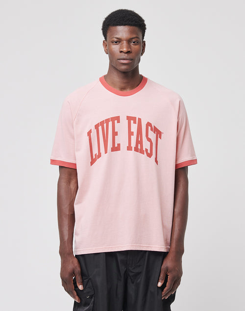 A person faces forward, wearing the LIVE FAST DIE YOUNG Live Fast Contrast Tee—light pink with red trim and bold “LIVE FAST” text, featuring raglan sleeves and soft cotton—paired with black pants against a plain gray background.