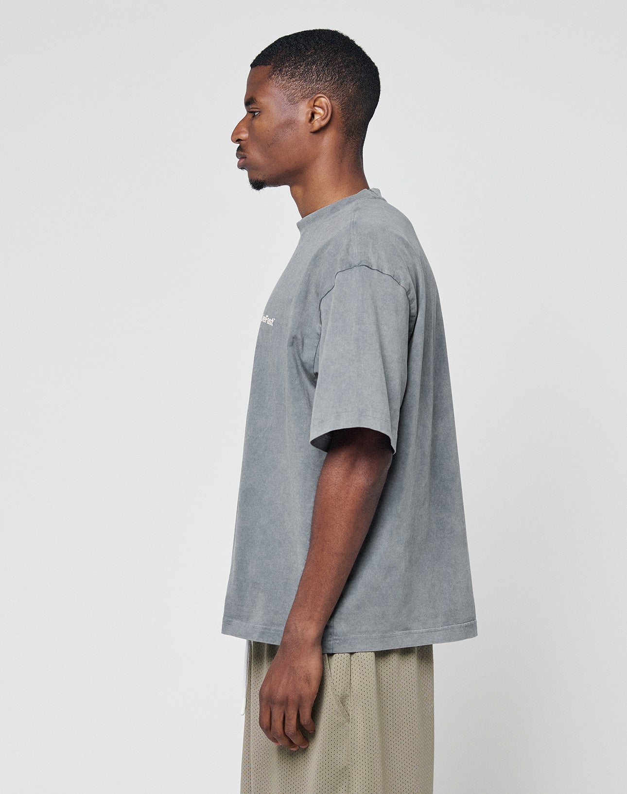 A man stands in profile against a plain background, wearing the LIVE FAST DIE YOUNG Basic +49 Chest Tee in gray with an oversize fit and 100% Baumwolle, paired with beige mesh shorts.
