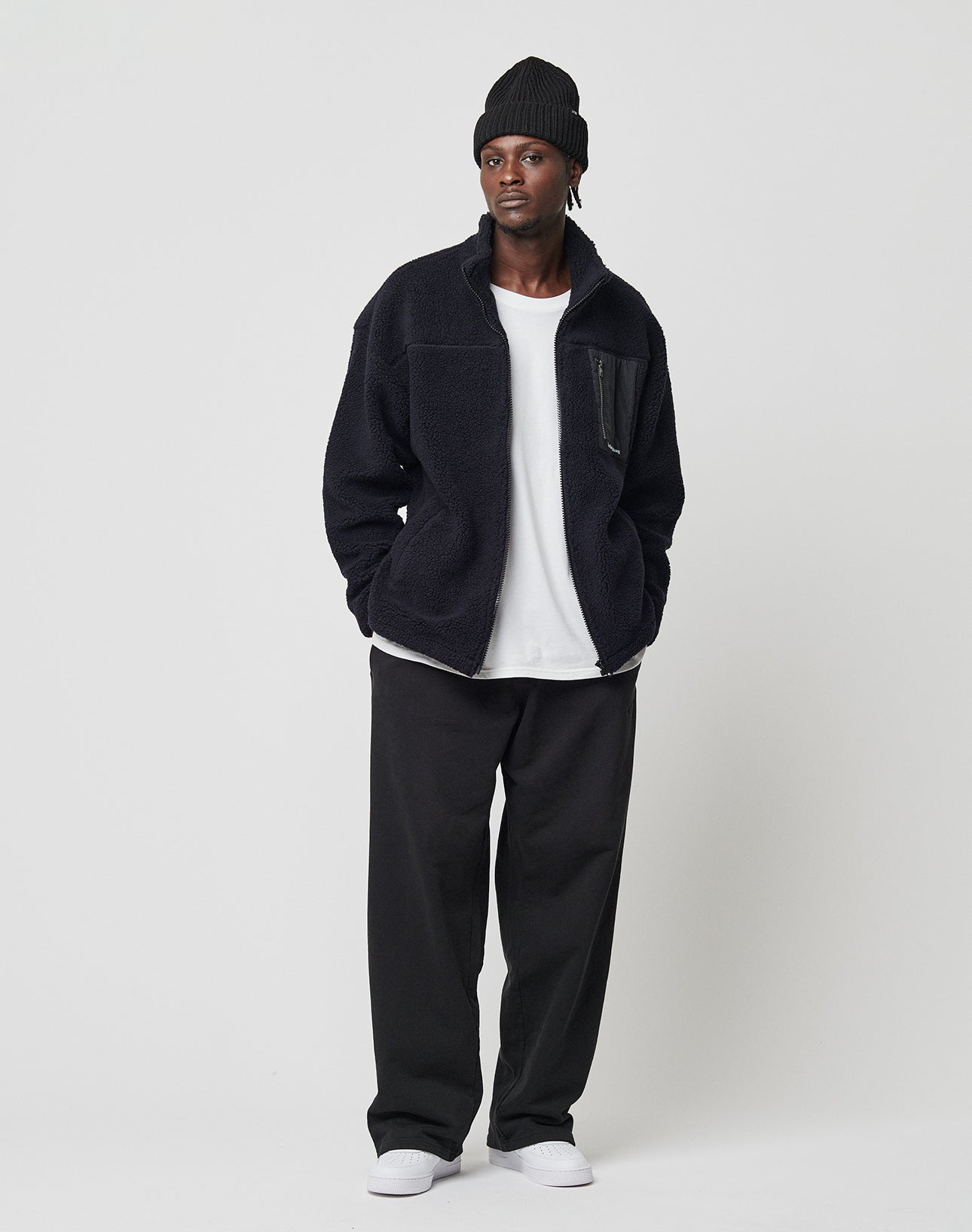 A person wearing the LIVE FAST DIE YOUNG Basic Teddy Jacket, a black beanie, white t-shirt, loose black pants, and white shoes stands against a plain light background with hands in pockets and a relaxed expression.