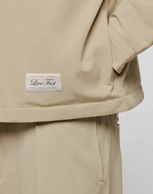 Close-up of a person wearing beige pants and the LIVE FAST DIE YOUNG LFDY Padded Shirt, an oversize fit with brusttaschen and a white “Live Fast” rectangular patch on the lower edge.