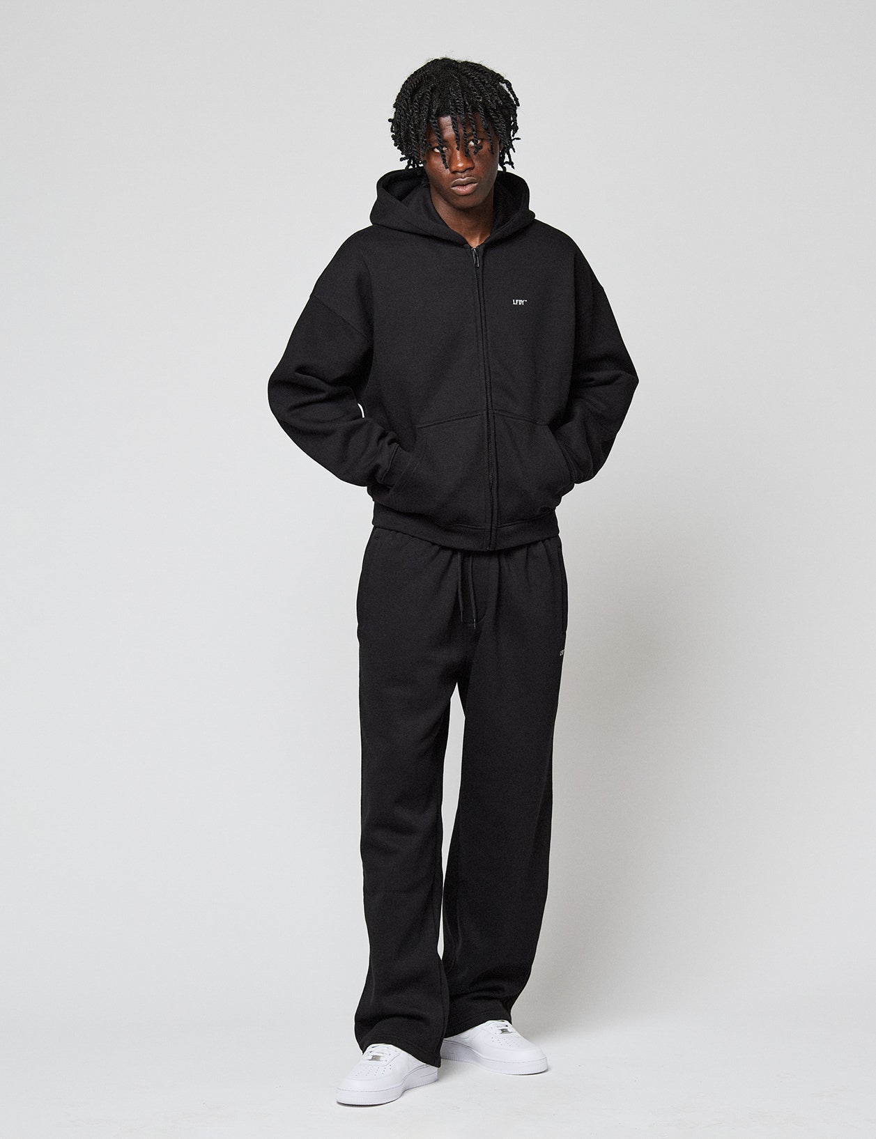 A person with short, curly hair wears the LIVE FAST DIE YOUNG Basic Hooded Zip and matching black sweatpants, standing hands-in-pockets against a plain white background with white sneakers.