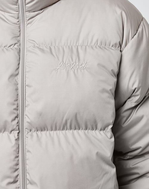 A close-up of the LFDY Puffer Jacket by LIVE FAST DIE YOUNG in light gray, showcasing its oversize fit, visible front zipper, and embroidered white logo stitching on the chest.