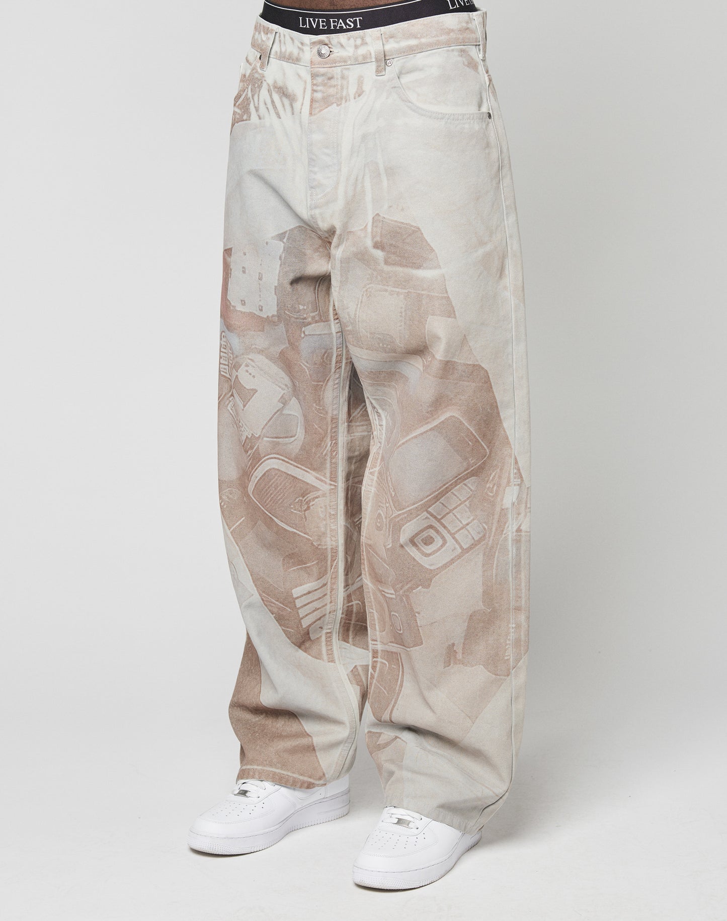 Someone wears the LIVE FAST DIE YOUNG LFDY Stolen Phones Denimâbaggy, light-colored jeans with a faded abstract electronics printâpaired with white sneakers and black LIVE FAST underwear visible above the waistband.