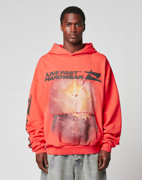 A man wears the LFDY Angel Hooded by LIVE FAST DIE YOUNG—an oversize red hoodie with abstract graphics, LIVE FAST HARDWEAR print, and a subtle distressed effect—standing against a plain light gray background.