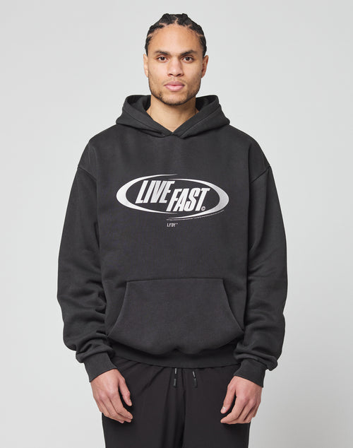A person in an oversize black LFDY Sphere Hooded by LIVE FAST DIE YOUNG, featuring a bold silver LIVE FAST. oval front print, stands facing forward against a plain light gray background.