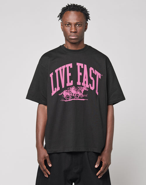 Wearing the Live Fast Stallion Tee by LIVE FAST DIE YOUNG—a black oversize 100% Baumwolle shirt with bold pink “LIVE FAST” frontprint above two horses—the person stands against a plain light gray background, facing forward neutrally.