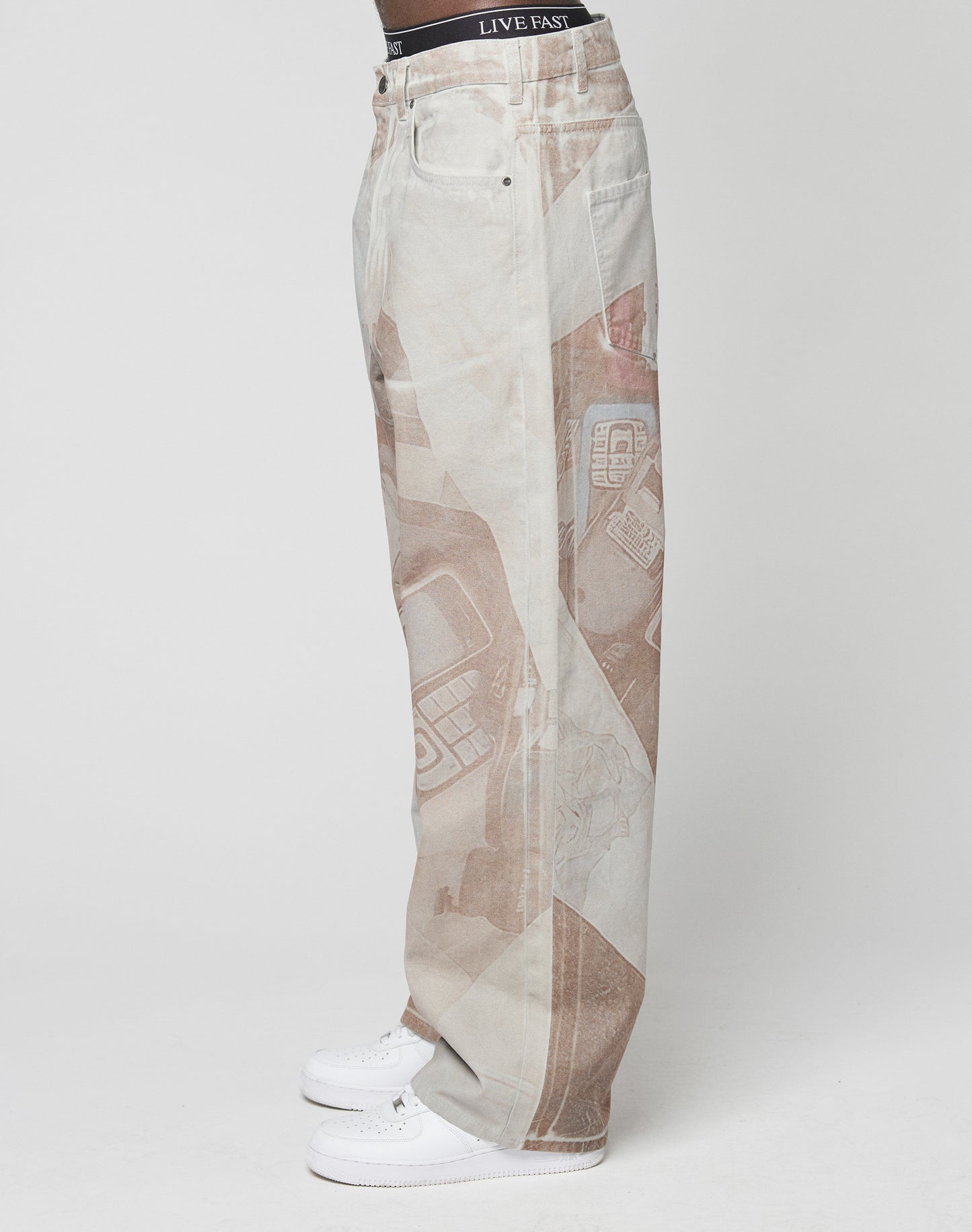 A person stands in profile against a white background, wearing LIVE FAST DIE YOUNGâs LFDY Stolen Phones Denimâbaggy, light-wash jeans with abstract brown printsâand white sneakers. The black LIVE FAST waistband peeks above the jeans.