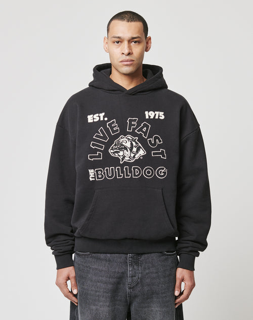 A person in a black LFDY The Bulldog Hooded by LIVE FAST DIE YOUNG, featuring an embroidered EST. 1975 LIVE FAST THE BULLDOG and bulldog graphic, paired with dark jeans, stands against a plain white background.