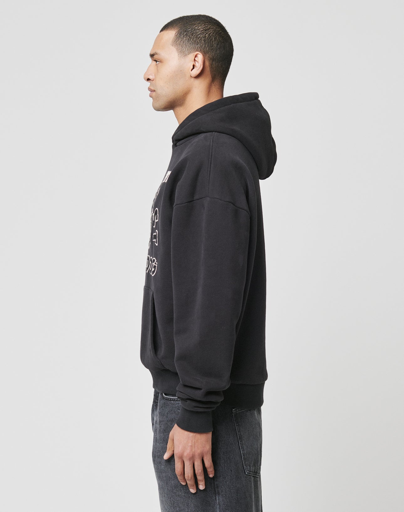 A man stands in profile facing left, wearing the LIVE FAST DIE YOUNG LFDY The Bulldog Hooded in black and dark jeans, against a plain light gray background.