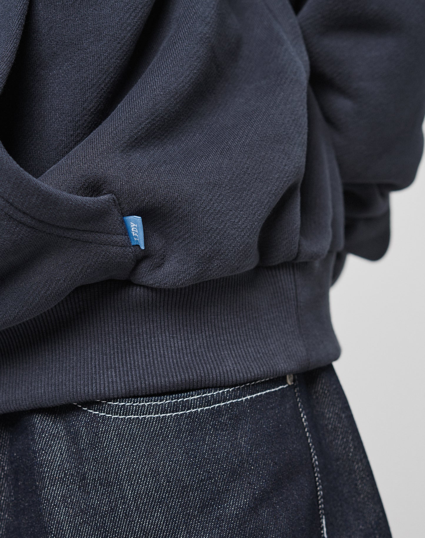 Close-up of a person wearing the LIVE FAST DIE YOUNG LFDY Script Hooded sweatshirt in dark navy waffle fabric with a blue tag, paired with dark denim jeans with white stitching. Only the lower oversize-fit sweatshirt and jean waistband are visible.