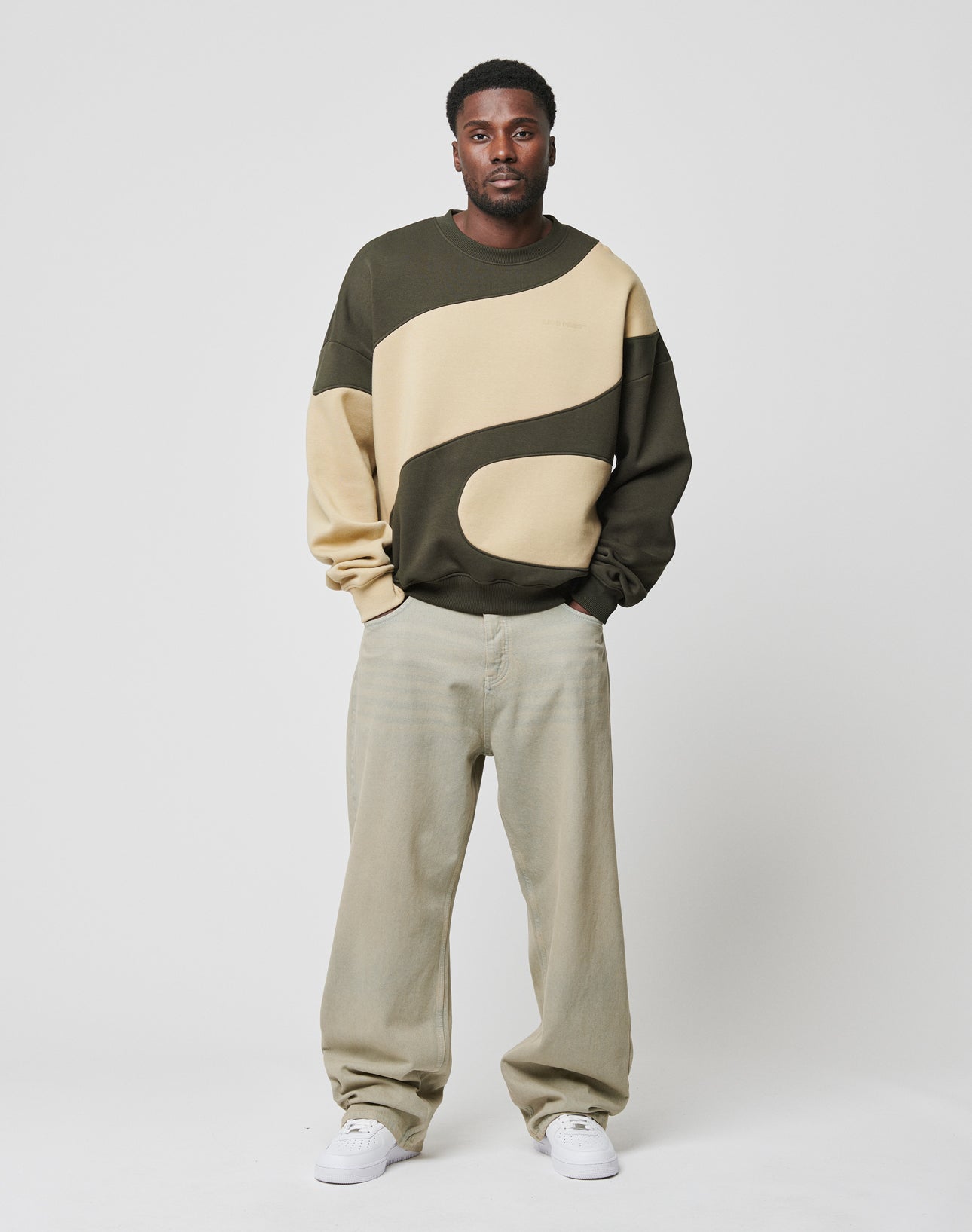A man stands against a white background wearing the LIVE FAST DIE YOUNG LFDY Wavy Sweater in dark green and beige, paired with light beige baggy pants and white sneakers, with his hands in his pockets.
