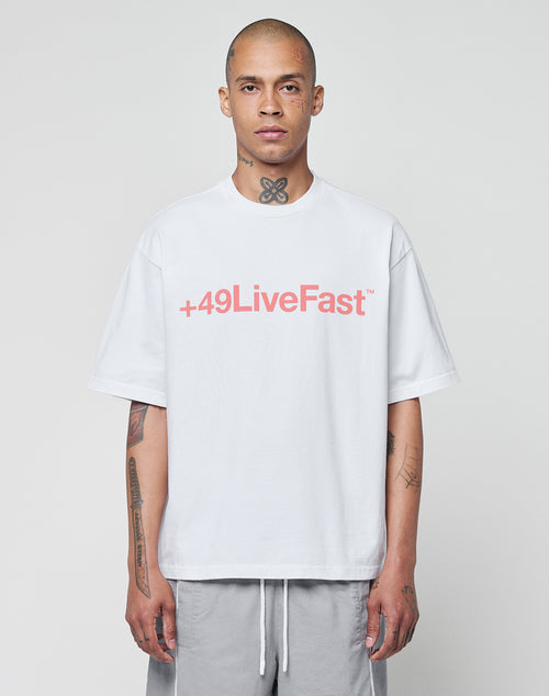 A person with a shaved head faces forward against a plain light background, wearing the LIVE FAST DIE YOUNG Basic +49 Tee—an oversized white cotton shirt featuring a red +49LiveFast front print—paired with gray shorts and showing neck and arm tattoos.