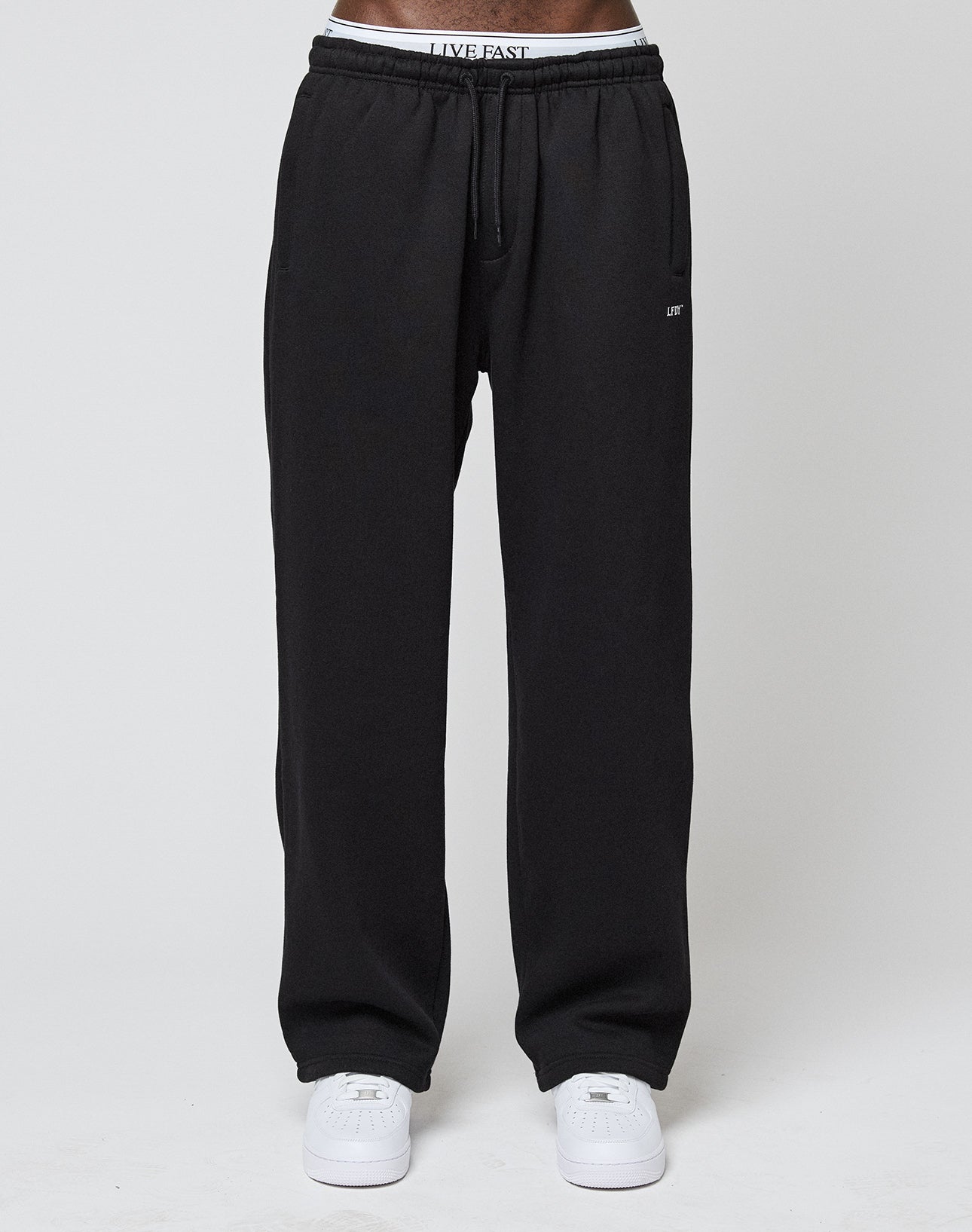 A person wears LIVE FAST DIE YOUNG Basic Wide Jogger sweatpants with an elastic waistband and logo print, partially revealing white underwear with LIVE FAST lettering, plus white sneakers, standing against a plain light background.