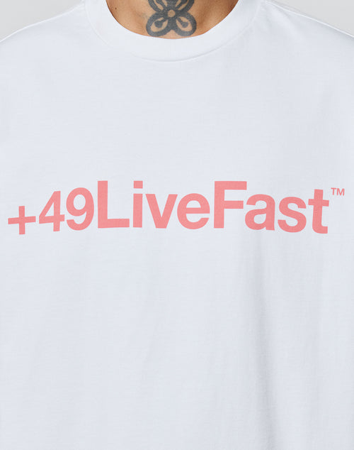 Someone wears the LIVE FAST DIE YOUNG Basic +49 Tee—an oversized white shirt with a bold pink +49LiveFast™ front print, crafted from soft Baumwolle for comfort. A floral or abstract neck tattoo shows above the collar.