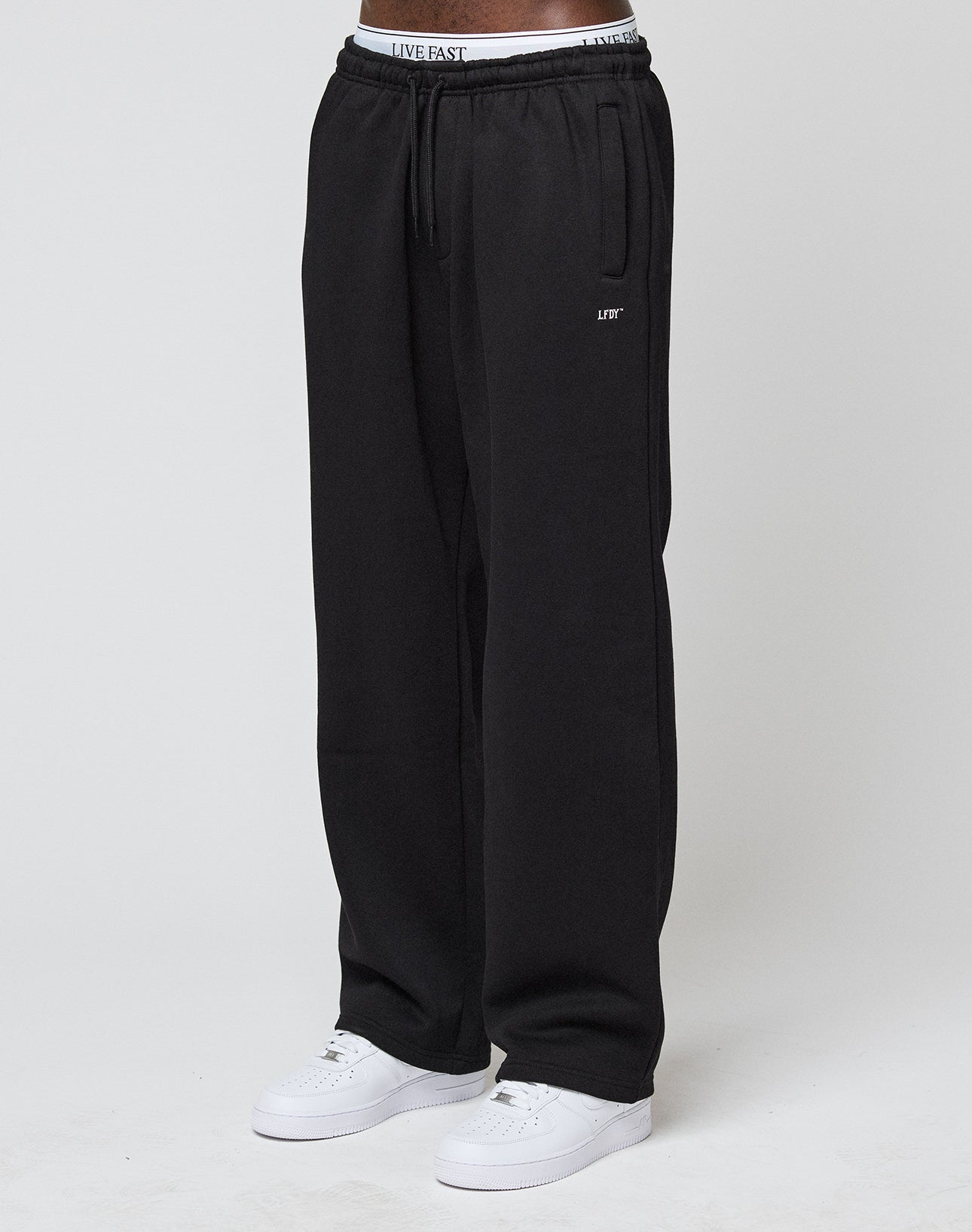 A person stands against a plain white background, wearing LIVE FAST DIE YOUNGs Basic Wide Jogger in black (loose fit), white sneakers, and a visible white waistband with the LIVE FAST logo.