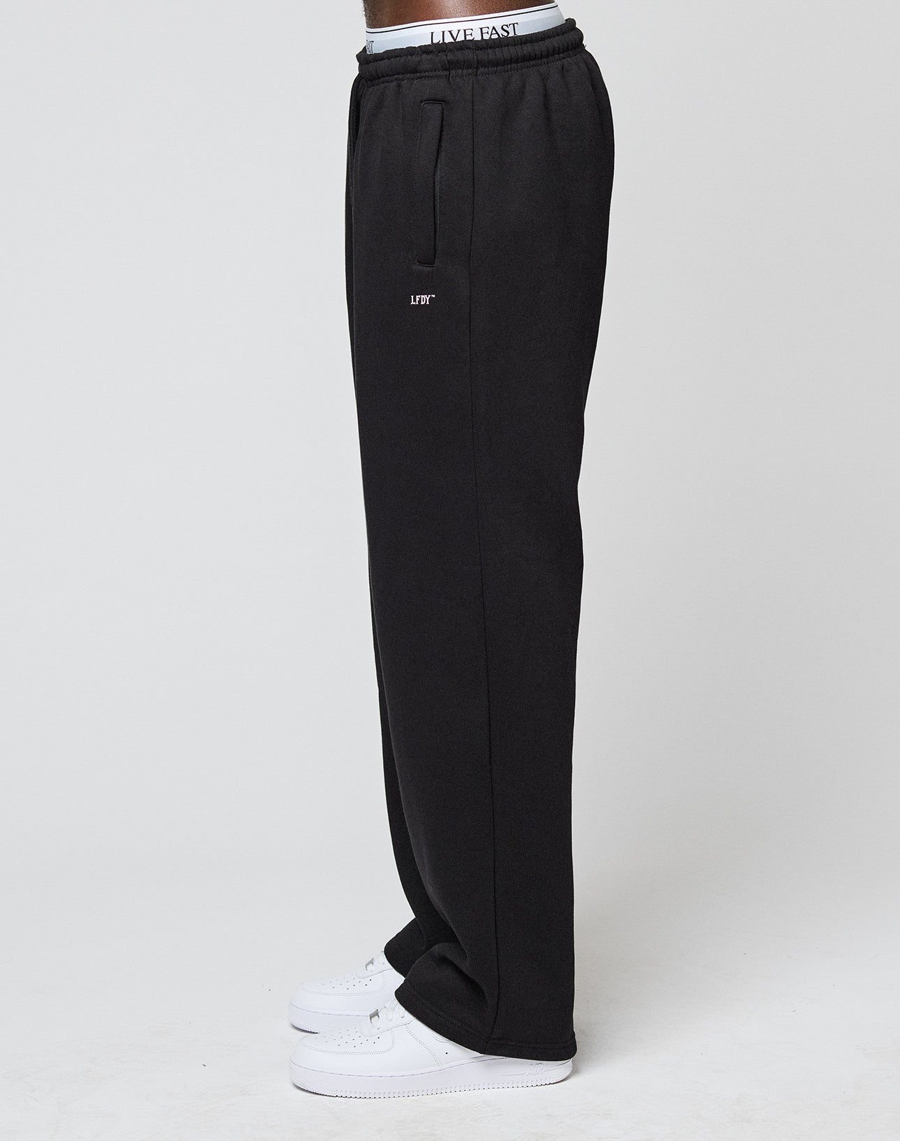 A person stands sideways against a plain light background wearing LIVE FAST DIE YOUNG Basic Wide Jogger black sweatpants with LIFT logo on the thigh, a white LIVE FAST elastic waistband, and white sneakers.