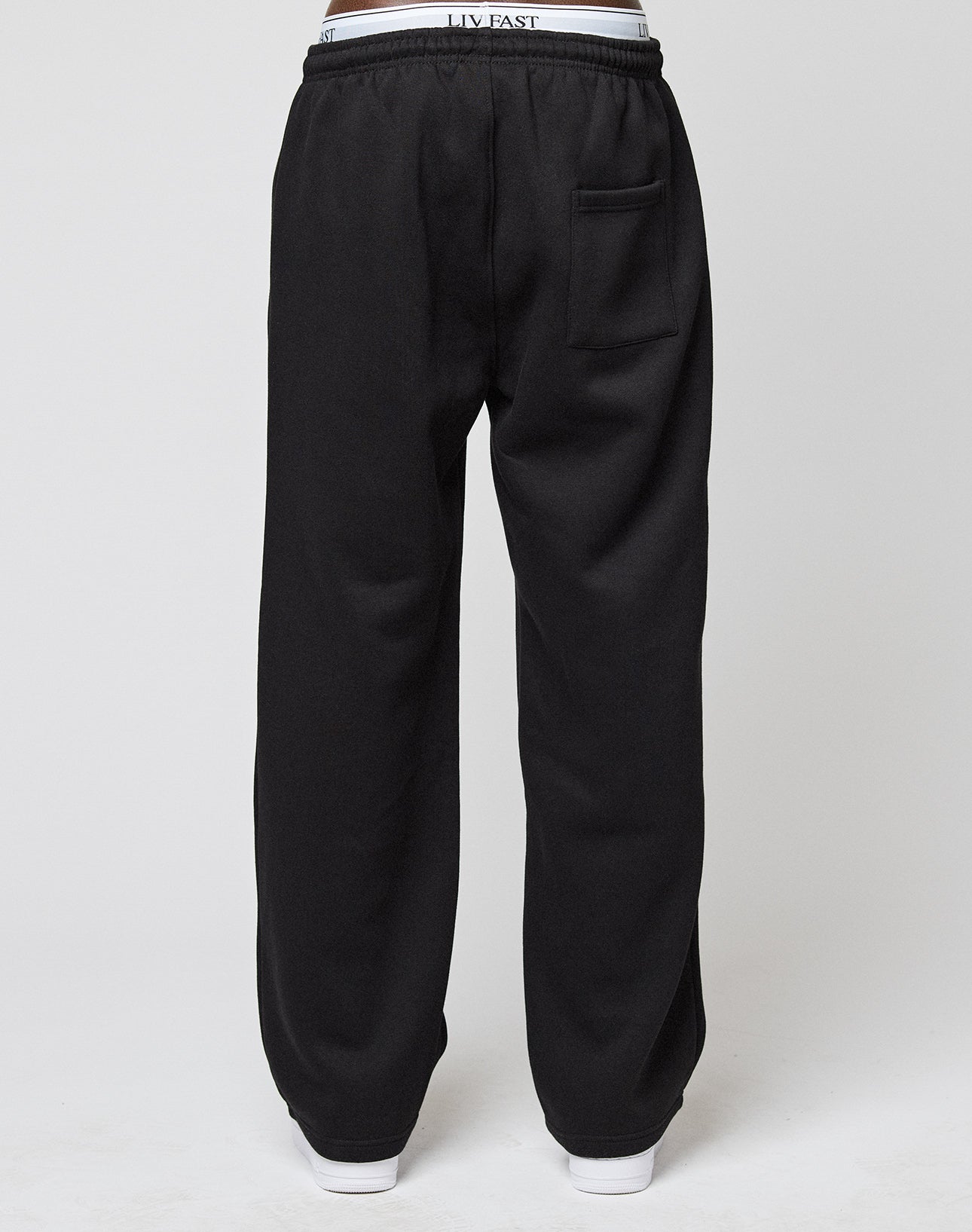 A person is shown from the back wearing LIVE FAST DIE YOUNG Basic Wide Jogger sweatpants in black with an oversize fit, elastic waistband, back pocket, white visible waistband, and white shoes against a plain light gray background.
