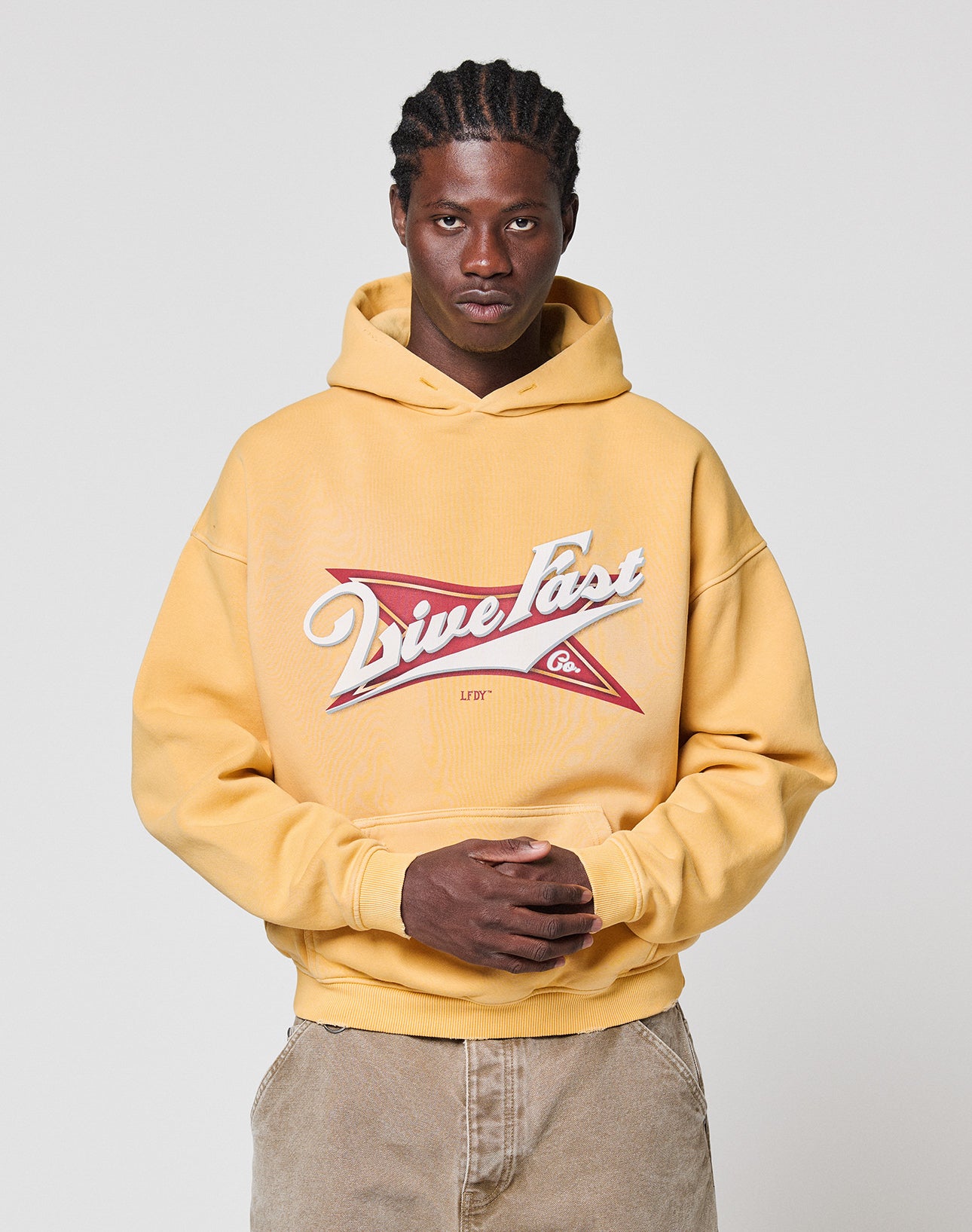 A man stands against a plain background wearing the LFDY Corp. Hooded by LIVE FAST DIE YOUNG, featuring a bold "Live Fast" front print in an oversize fit, paired with tan pants. His hands are clasped as he looks directly at the camera.