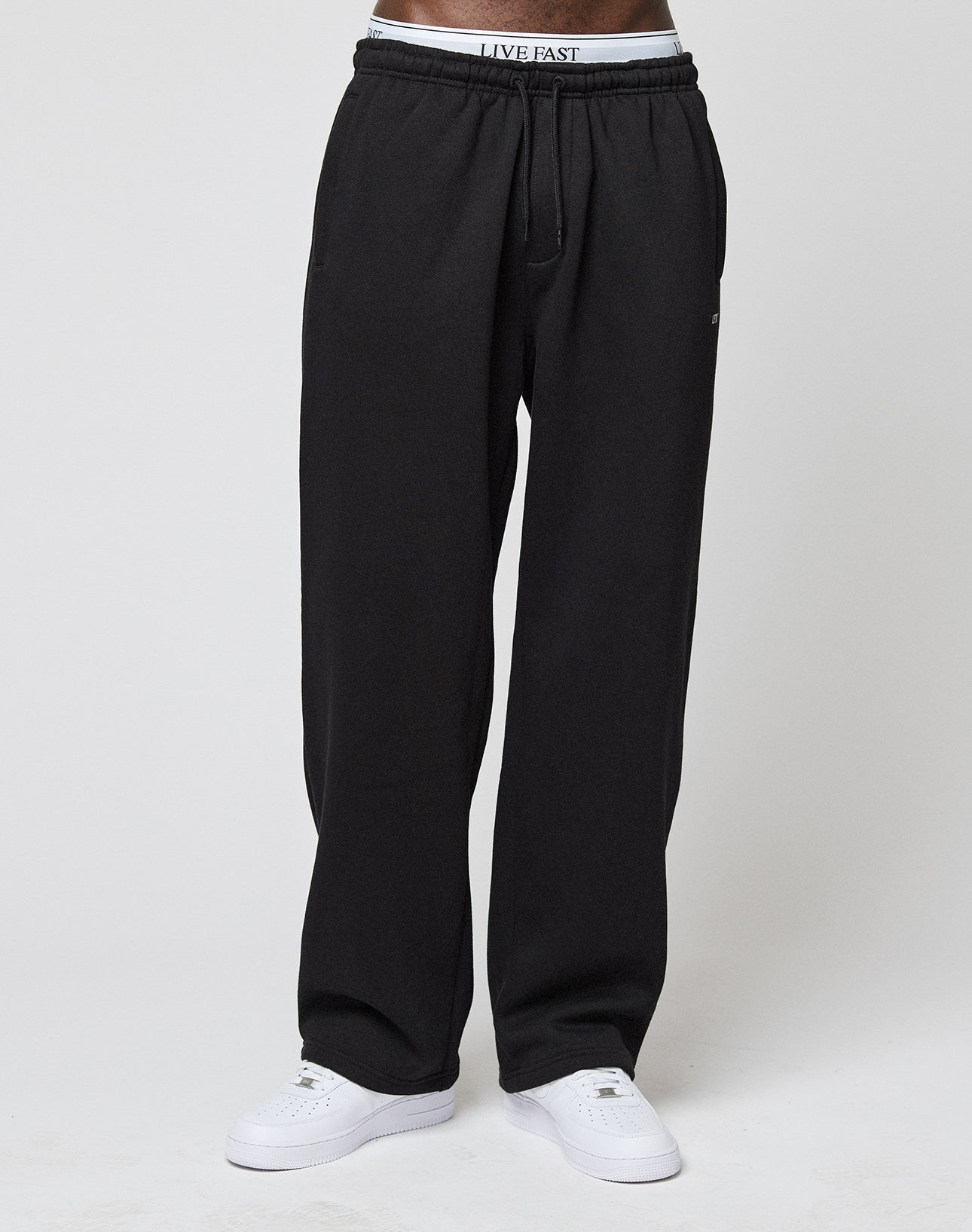 A person wears Basic Wide Jogger sweatpants by LIVE FAST DIE YOUNG, paired with white sneakers and logo waistband underwear reading LIVE FAST, standing against a plain white background.