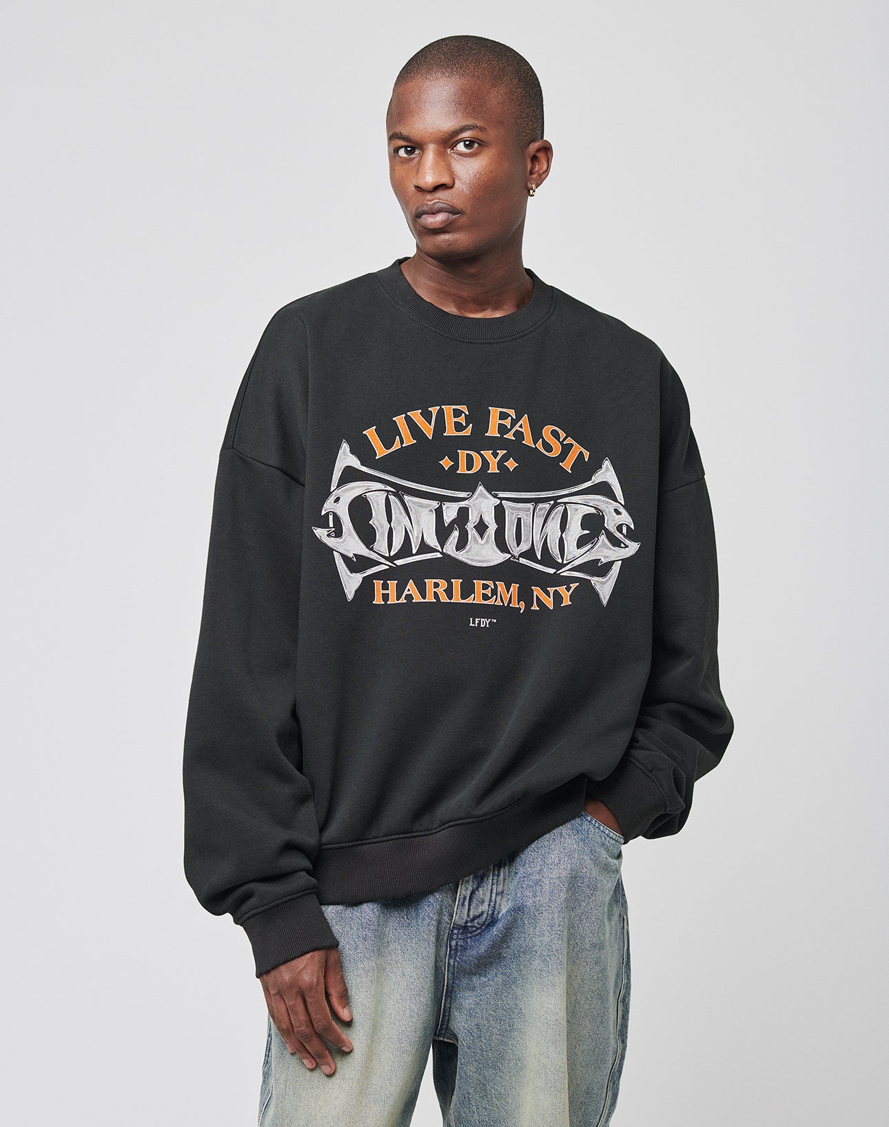 A person wears the Live Fast Tribal Sweater by LIVE FAST DIE YOUNG, an oversized black cotton-polyester sweatshirt with tan and white HARLEM, NY graphics, paired with light blue jeans against a simple light background.