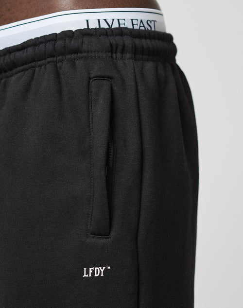 Close-up of a person wearing the LIVE FAST DIE YOUNG Basic Wide Jogger in black, featuring a white embroidered LFDY logo near the pocket and an elastic waistband, styled with white underwear printed LIVE FAST above the pants.
