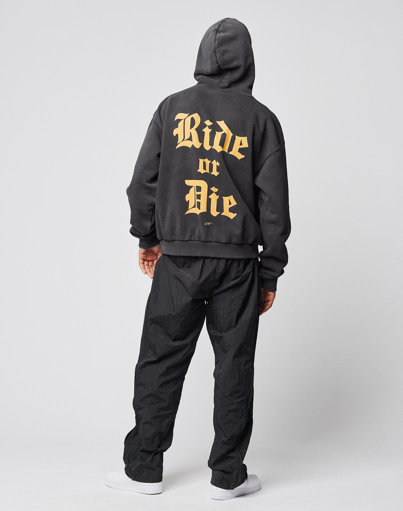 A person faces away, wearing the LIVE FAST DIE YOUNG Ride or Die Hooded—a black oversized hoodie with double-layer hood and large beige gothic Ride or Die print on the back—paired with black pants and white shoes against a plain background.