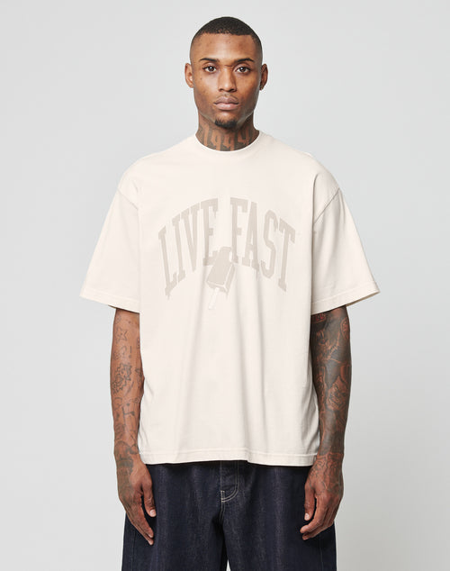 A tattooed man wears the LIVE FAST DIE YOUNG Live Fast Ice Tee, a loose beige Baumwolle t-shirt in Oversize Fit with a LIVE FAST print and popsicle graphic. He stands facing forward against a plain gray background, arms relaxed.