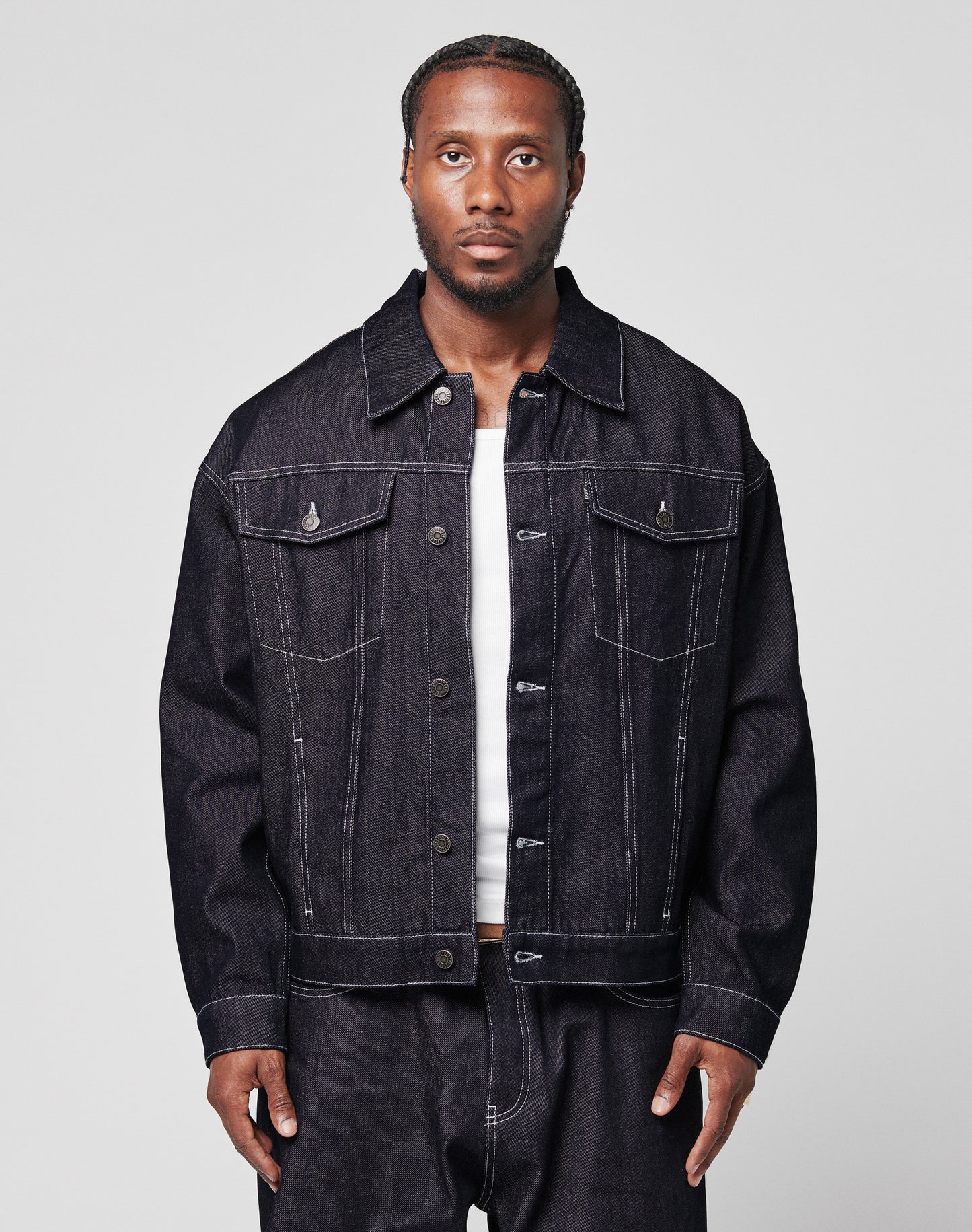 A man with braided hair wears an oversize LFDY Denim Jacket by LIVE FAST DIE YOUNG and matching raw-denim jeans over a white shirt, standing against a plain light gray background.