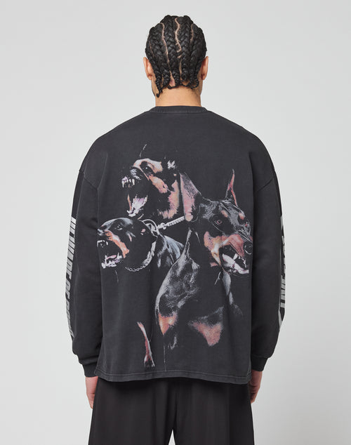 A person with braided hair faces away, wearing the LIVE FAST DIE YOUNG LFDY Canine Longsleeve—an oversize-fit black shirt made from 100% cotton, featuring three barking Dobermans on the back and light stripes on the sleeves.