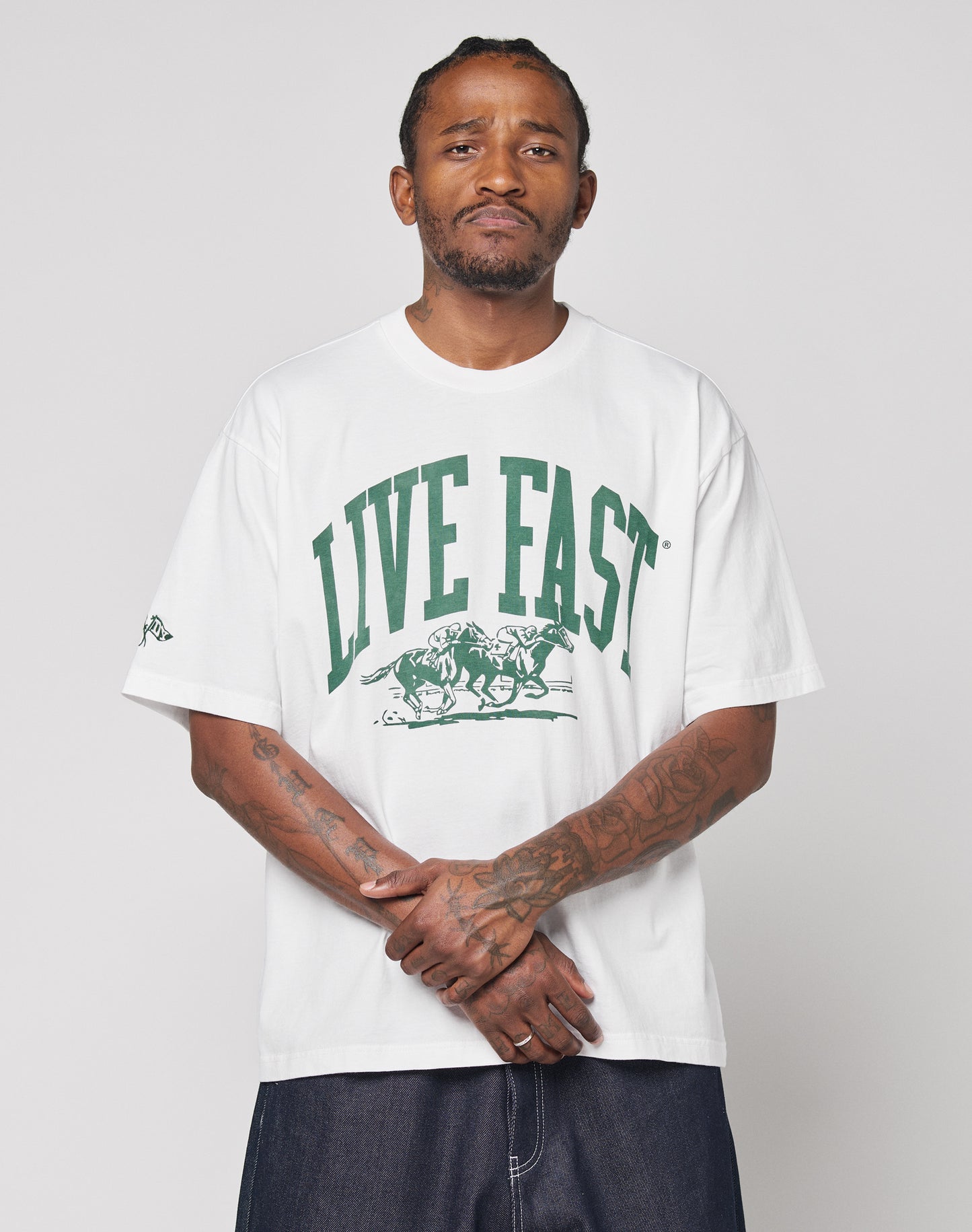 A tattooed man faces the camera wearing the Live Fast Stallion Tee by LIVE FAST DIE YOUNG, an oversize white 100% Baumwolle T-shirt with a green LIVE FAST and running horses print, paired with dark pants against a plain light background.