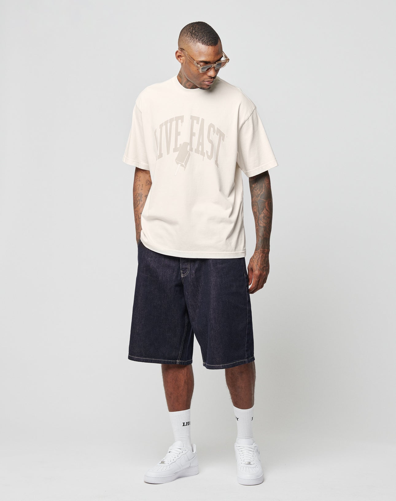 A man wears the LIVE FAST DIE YOUNG LFDY Jorts with a cream Flag-Label tee, white socks, and sneakers. He stands against a light gray background, looking down to his left with hands in pockets.