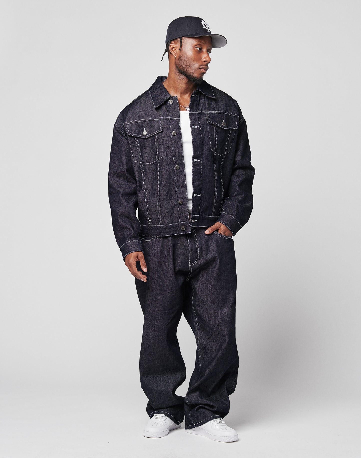 A man in an LFDY Denim Jacket by LIVE FAST DIE YOUNG, matching baggy jeans, a white T-shirt, white sneakers, and a dark baseball cap stands against a plain light background, looking to his left with a relaxed posture.