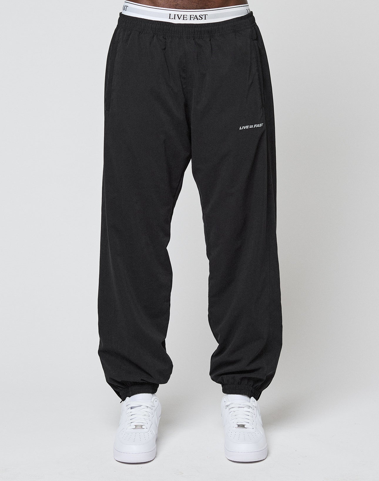 A person in regular fit LFDY Trackpants by LIVE FAST DIE YOUNG, featuring black polyester fabric and LIVE FAST text on the elastic waistband and near the left pocket, pairs them with white sneakers against a plain light background.