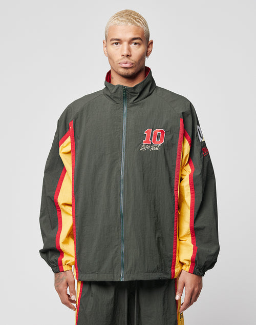 A person with short blond hair wears a LIVE FAST DIE YOUNG Westend Trackjacket in dark green, red, and yellow polyester with multi-logo embroidery, standing against a plain light gray background.