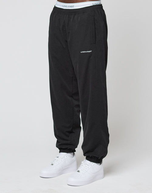 A person wearing LFDY Trackpants by LIVE FAST DIE YOUNG, featuring an elastic waistband and LIVE FAST text on the waistband and thigh, paired with white sneakers, stands against a plain white background.