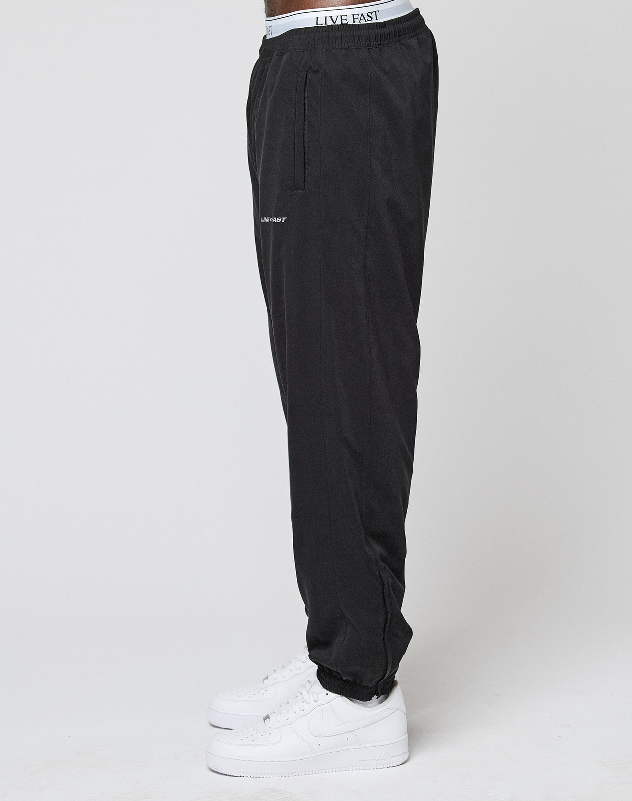 A person wearing LFDY Trackpants by LIVE FAST DIE YOUNG—regular fit, black polyester, with LIVE FAST branding on the elastic waistband and left thigh—paired with white sneakers, stands sideways against a plain white background.