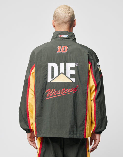 A person with short, bleached hair is seen from behind wearing the LIVE FAST DIE YOUNG Westend Trackjacket, a regular fit dark green polyester jacket with yellow and red stripes and a large Multi-Logo design reading 10, DIE, and Westend on the back.