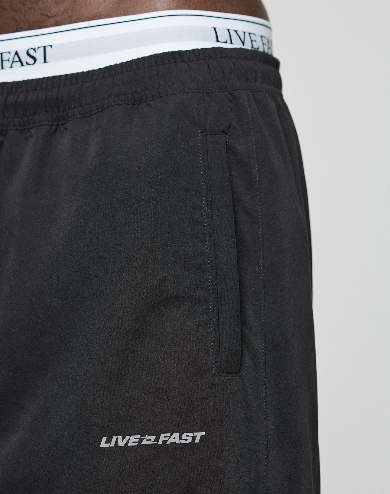 Close-up of a person wearing LFDY Trackpants by LIVE FAST DIE YOUNG. The black polyester shorts have a side pocket, Regular Fit, and feature LIVE FAST printed in white near the pocket plus an Elastikbund waistband with matching text.