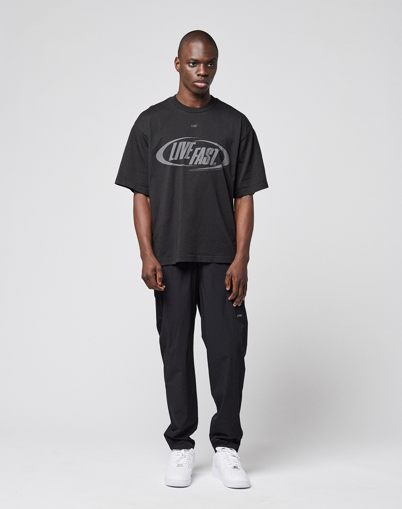 A man stands against a plain white background wearing the LIVE FAST DIE YOUNG LFDY Sphere Tee with front and back prints, black pants, and white sneakers. He faces forward neutrally with his arms by his sides.