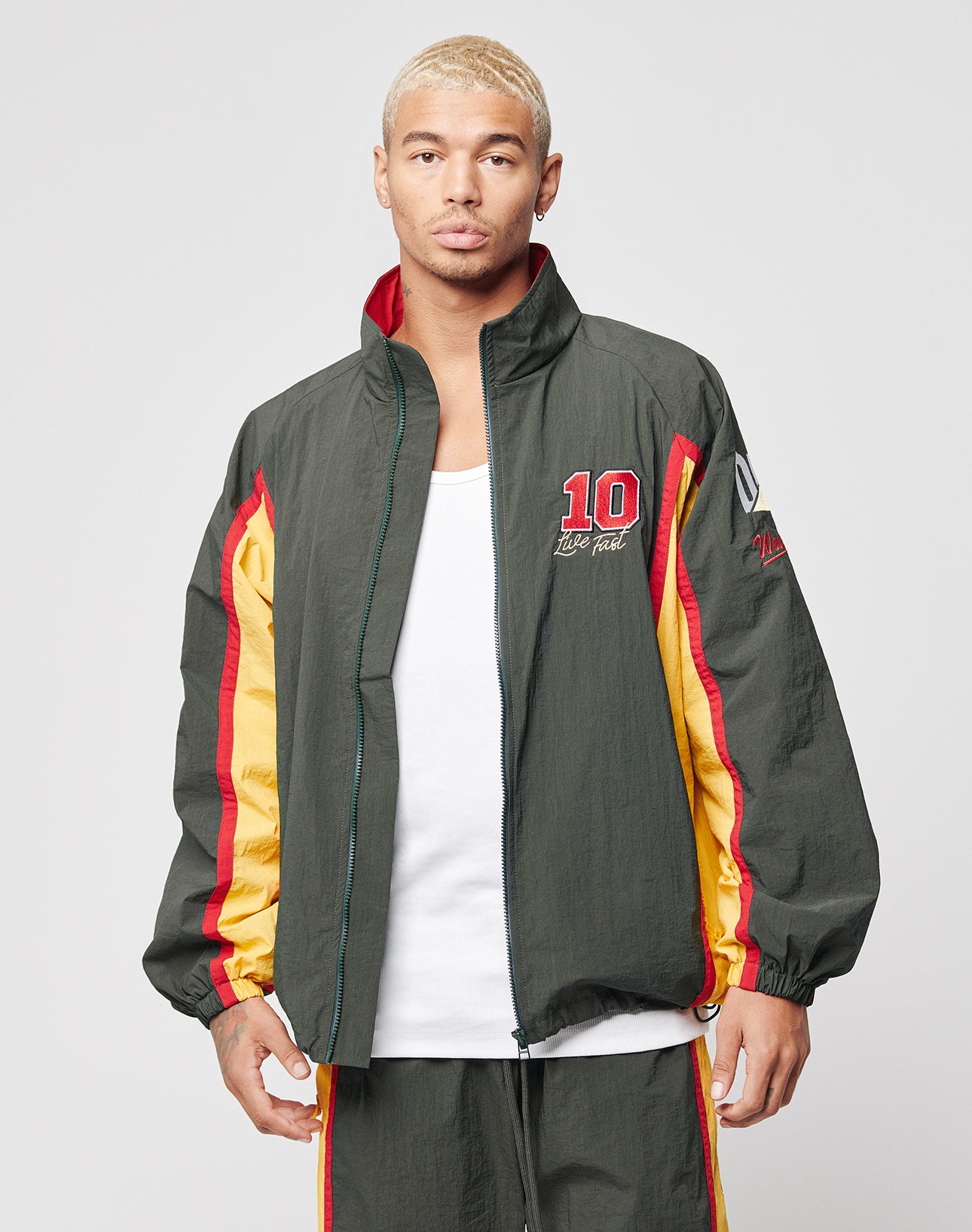 A man with short blond hair wears the LIVE FAST DIE YOUNG Westend Trackjacketāregular fit, multi-logo, in dark green, red, and yellow polyester with 10 on the chestāover a white tank, posing neutrally against a gray background.