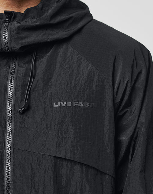 Close-up of the Live Fast Nylon Jacket by LIVE FAST DIE YOUNG, featuring a black, crinkled fabric with a hood and zipper. A gray LIVE FAST Silikon-Print appears on the chest. Only part of the wearer’s neck and jaw is visible. Oversized fit.