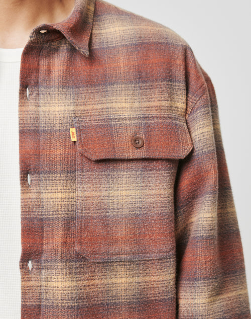 A person wears the LIVE FAST DIE YOUNG Vintage Padded Flannel in brown, rust, and beige plaid over a white textured shirt, highlighting its buttoned chest pocket and part of the collar.