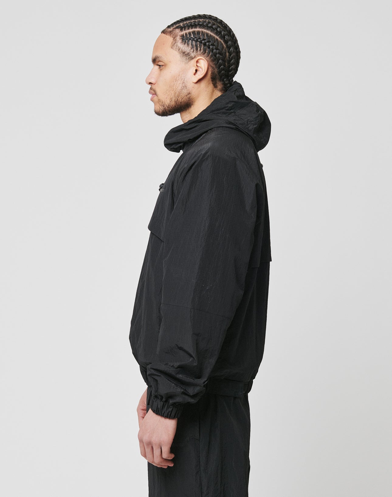 A man with braided hair stands in profile against a plain background, wearing the LIVE FAST DIE YOUNG Live Fast Nylon Jacket with seitliche Eingrifftaschen and matching oversized pants.